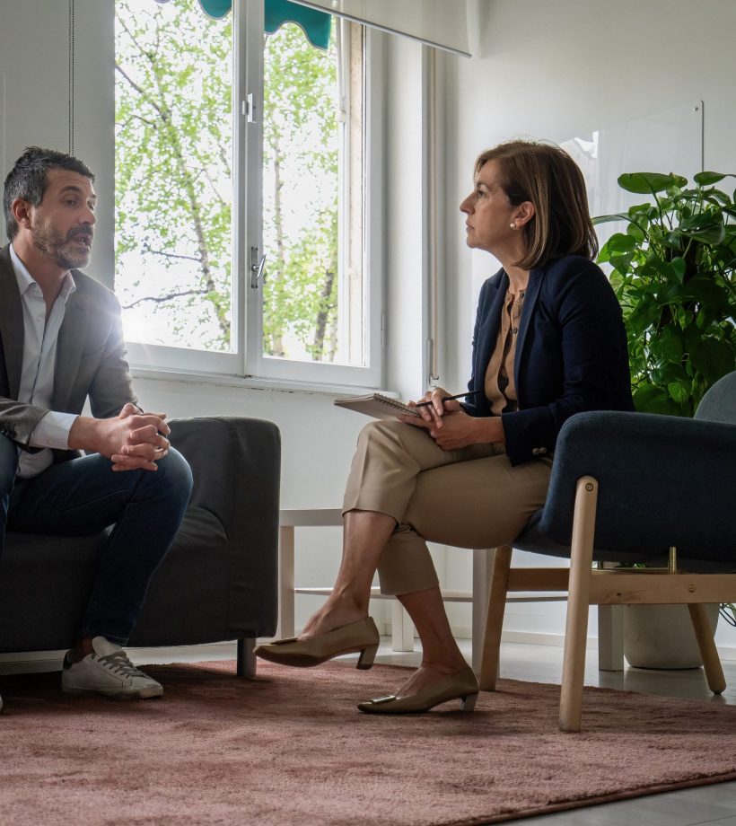 Psychotherapist with notebook conducting therapy session with male patient in bright, modern office. Natural light and plant create calming atmosphere for mental health counseling and support.